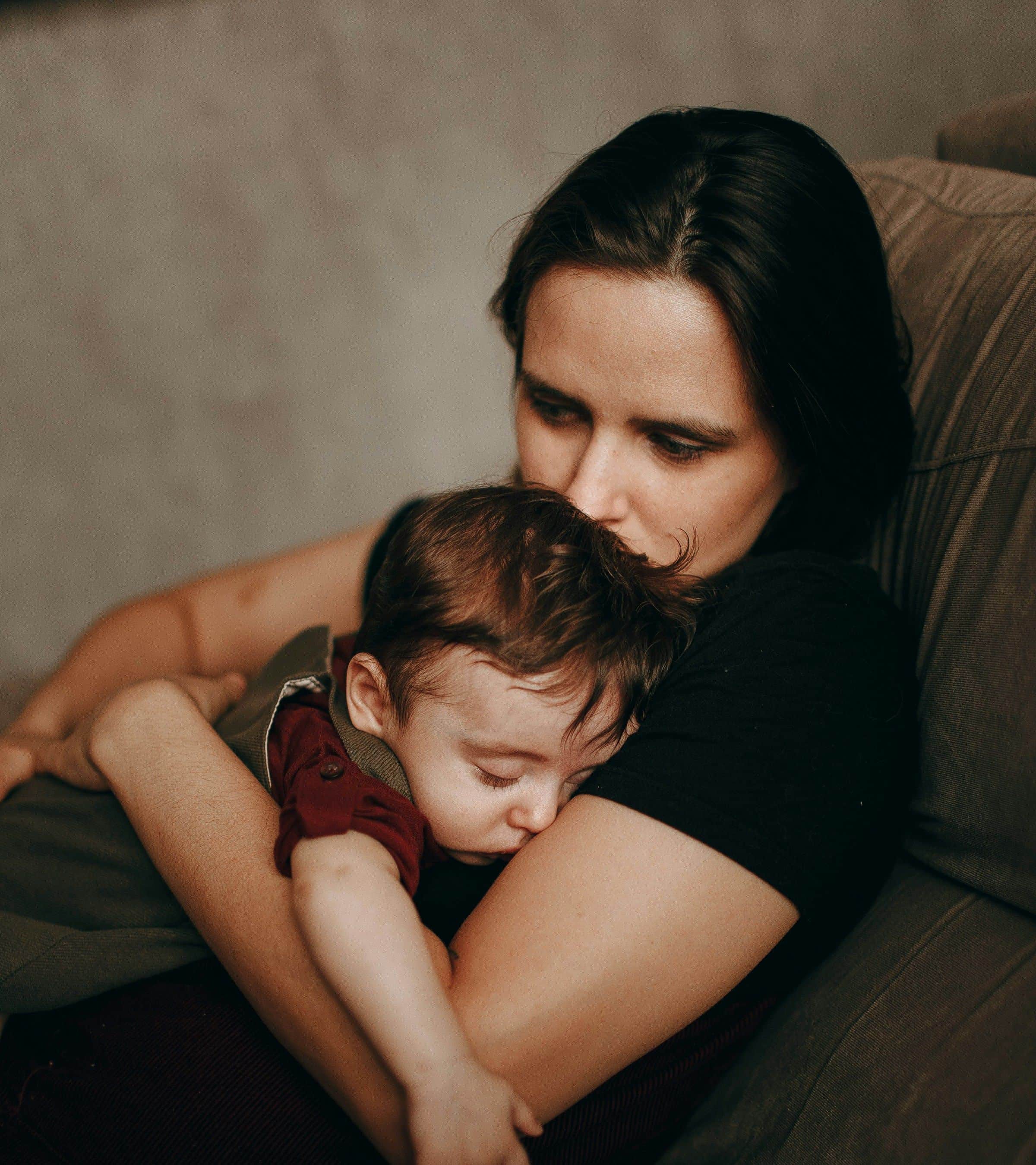 A mother on a sofa holds her sleeping baby close, her lips resting on his hair.