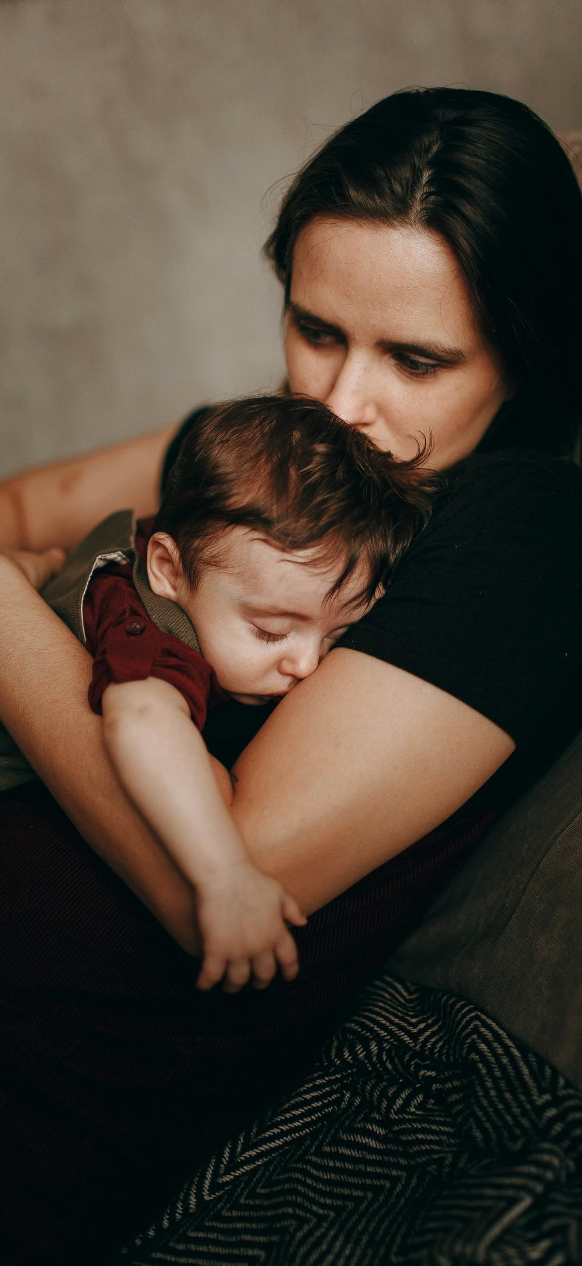 A mother on a sofa holds her sleeping baby close, her lips resting on his hair.